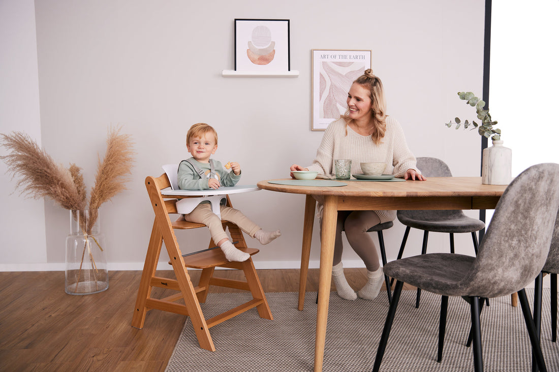 A baby sitting in a wooden high chair with a parent sitting near at a dining table