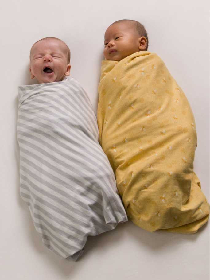 Two babies in Woolbabe swaddles lying on a white surface. One is yawning