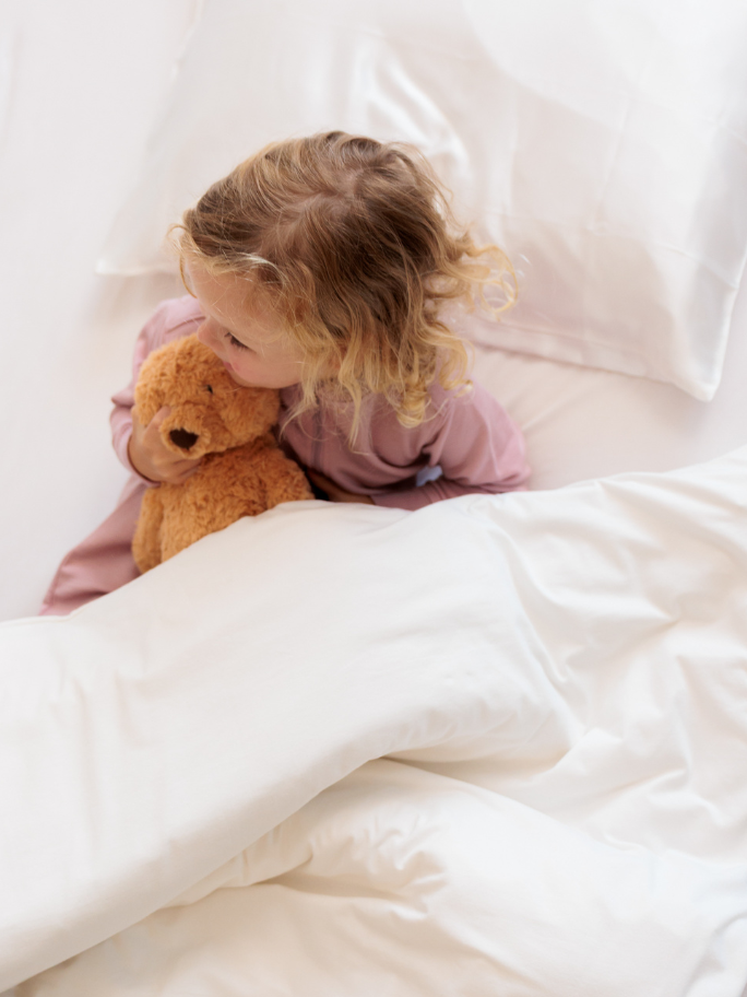 Toddler in white bedding and pink fleec pyjamas holding brown teddy