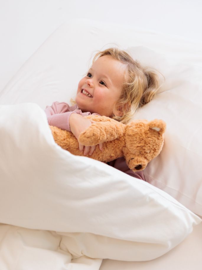 Toddler in white bedding, wearing fleece pyjamas and cuddling teddy