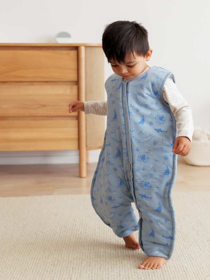 A toddler wearing a blue Woolbabe sleeping bag in a room setting