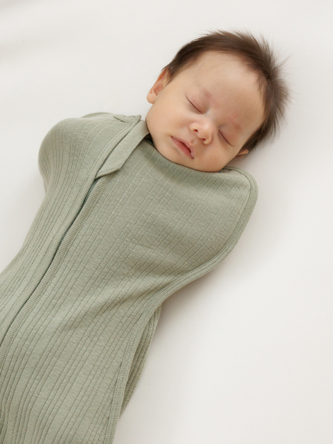 Newborn asleep wearing a green ribbed zip swaddle in a cot