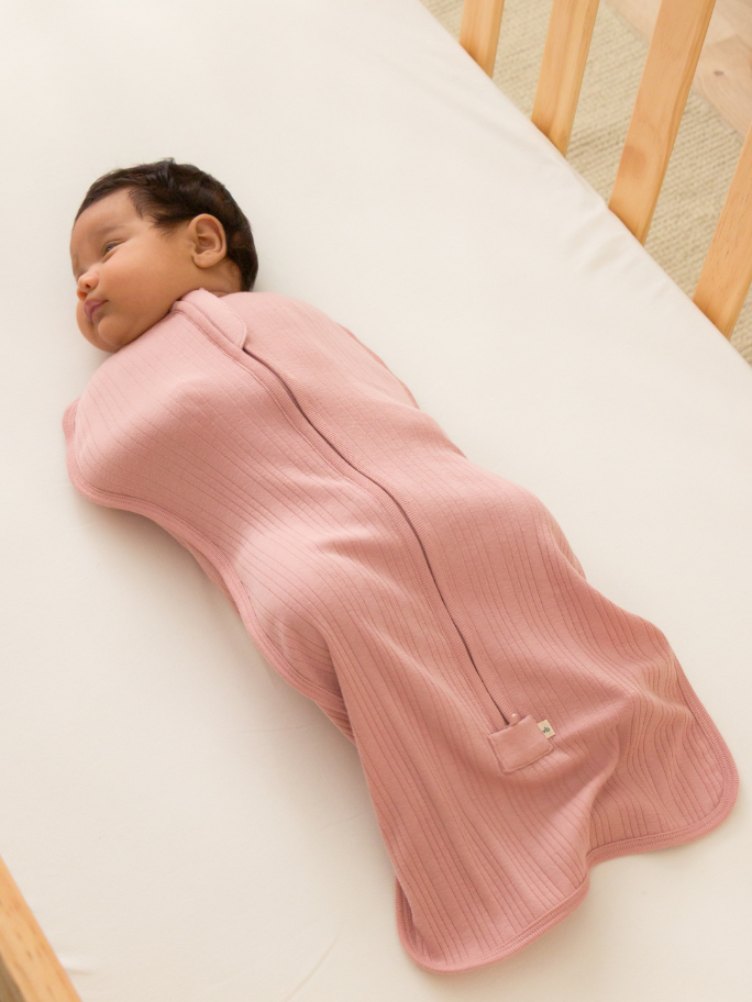 Baby in pink ribbed swaddle bag lying inside a cot with plain white sheet