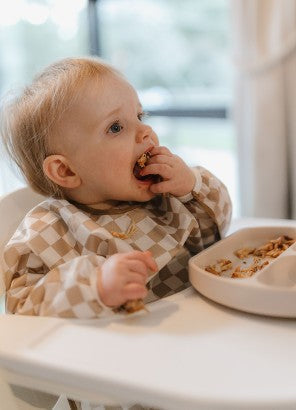 Baby eating food in a Hauck high chair with brown check sleeved bib