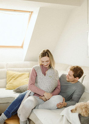 Father and mother sitting down on couch smiling at baby inside wrap