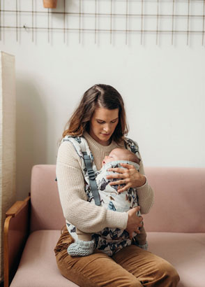 Mother sitting down on couch watching baby sleep inside front carrier