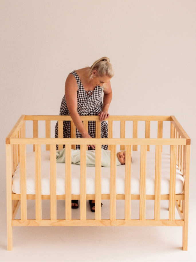 Mother gazing into Frank Cot at sleeping baby
