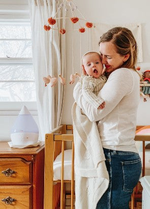Mum cuddling baby with Crane humidifier in background winter wellness kit