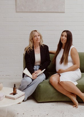 Two women sitting on a couch, one is pregnant. A coffee table on the side