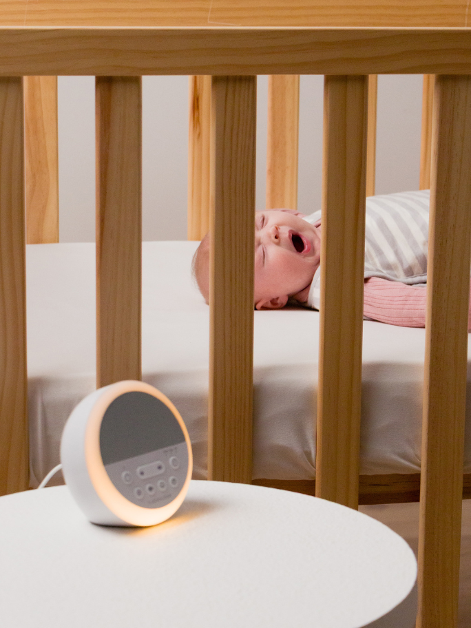 Baby in a cot, wearing a Woolbabe sleeping bag yawning with sound machine in the foreground