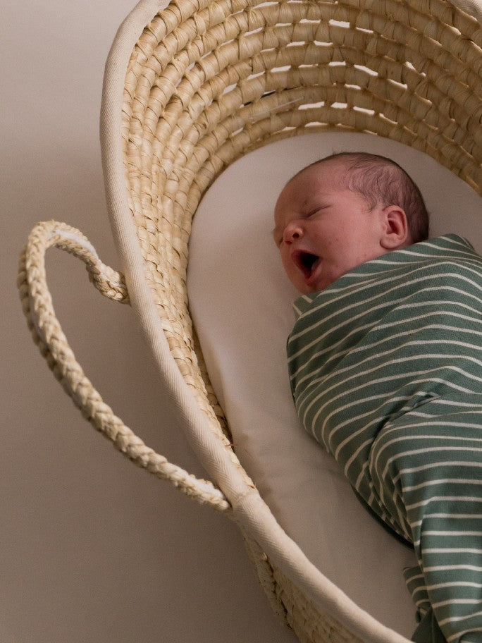 Baby in a green striped swaddle, yawning in a moses basket