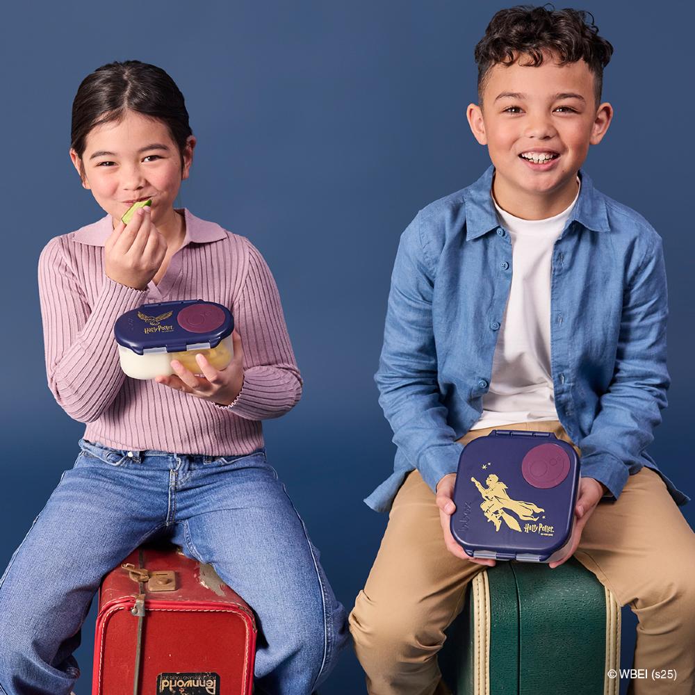 Two children sitting on suitcases with Harry Potter lunch boxes against a blue background