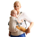 Woman carrying a baby in a beige cotton Beco Gemini baby carrier with a plain background
