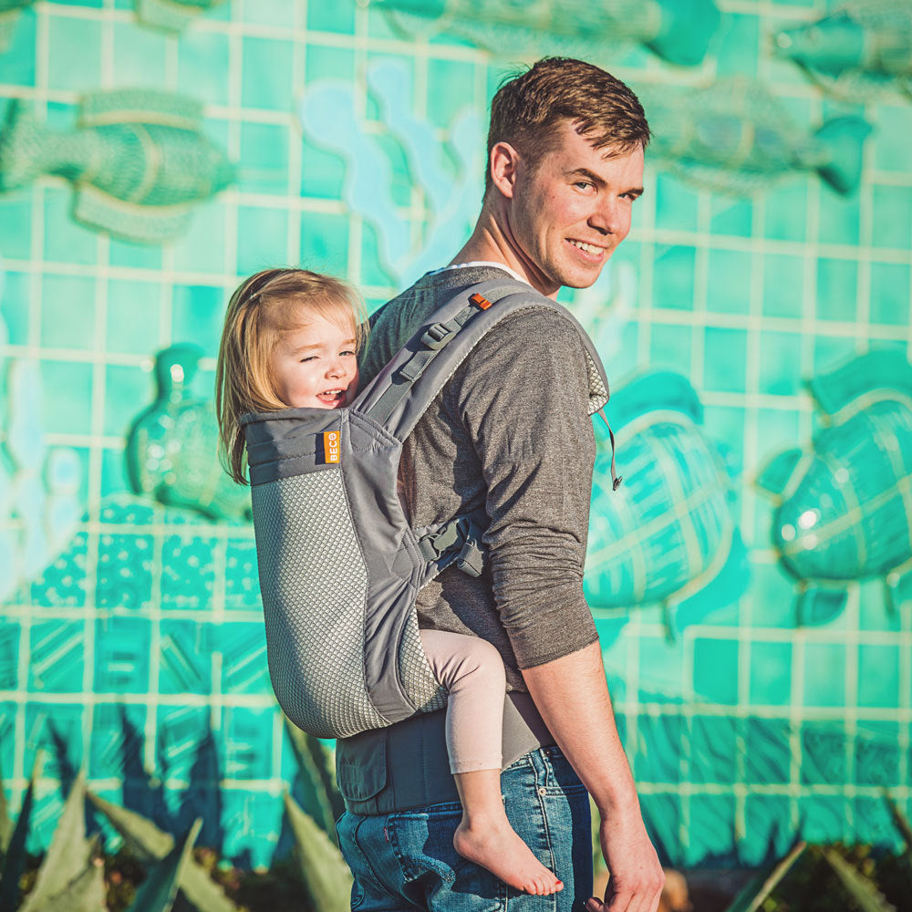 Man carrying a 4 year old child in a Beco Toddler carrier against a colorful tiled wall.