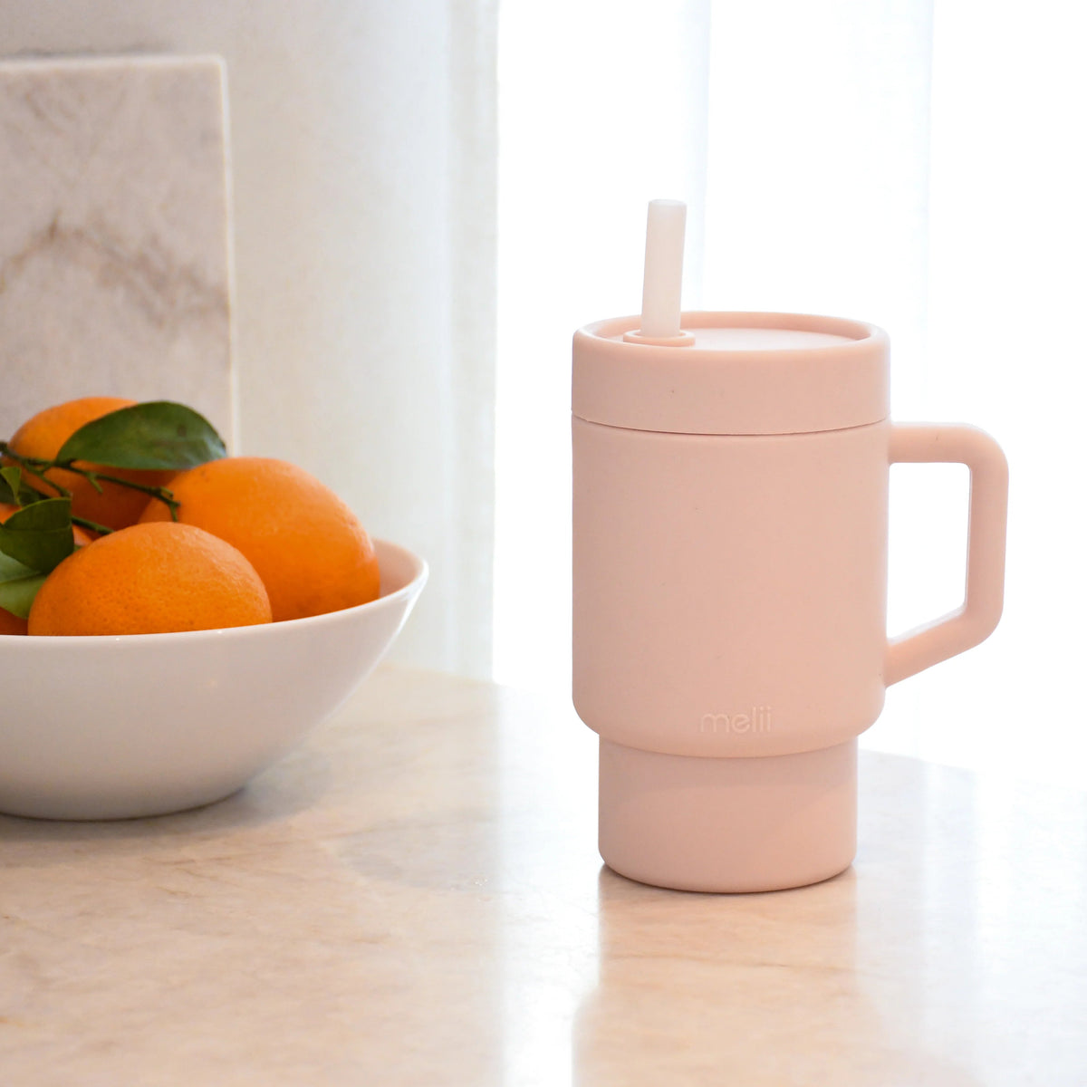 Pink silcione kids trainer tumblerug with a straw on a marble surface next to a bowl of oranges.
