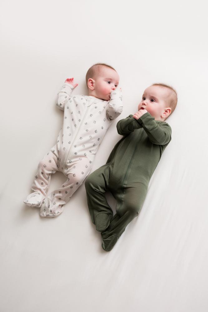 Two babies lying side by side on a white blanket, wearing footed sleepers. The baby on the left wears a light cream sleeper with small gray pattern dots, and the baby on the right wears an olive green sleeper.