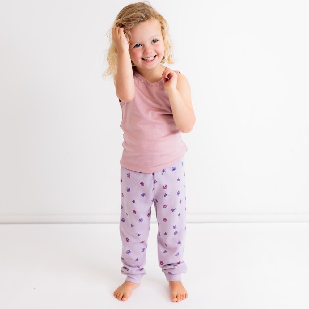 A young girl with blonde hair smiling and posing in a pink sleeveless top and light purple patterned pajama pants against a white studio background.