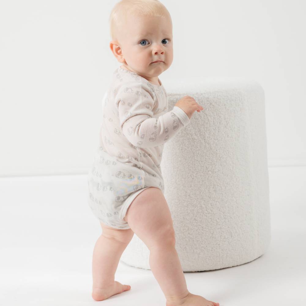Baby in a white onesie standing next to a white textured stool on a white background