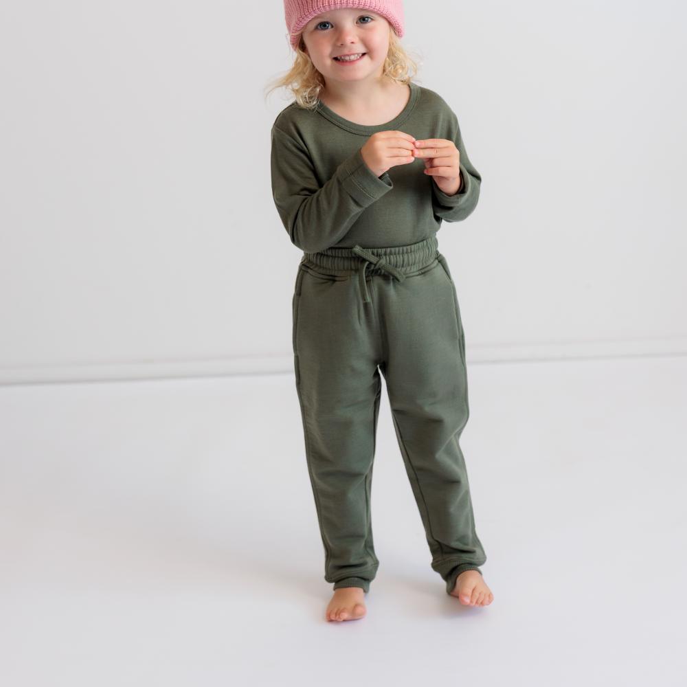 A toddler standing barefoot in a studio setting, wearing an olive green long-sleeve top and matching merino fleece jogger pants, with a pink knit hat.