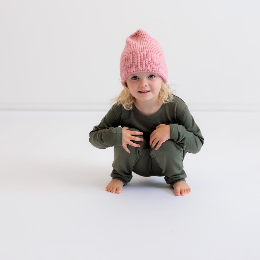 A toddler crouching on a white studio floor wearing an olive green long-sleeve pajama set and a pink knit beanie hat.