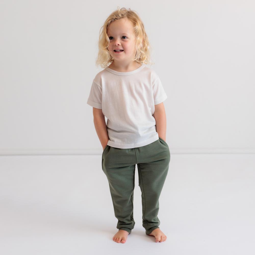 A young child with blonde curly hair stands barefoot against a plain white studio background, wearing a white short-sleeve t-shirt and olive green pants.