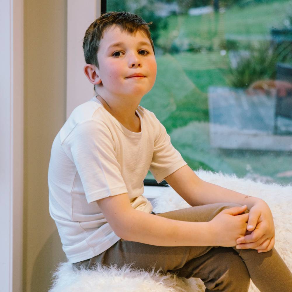 A young boy sitting on a white faux fur rug in front of a large window, wearing a  white t-shirt and brown pants.