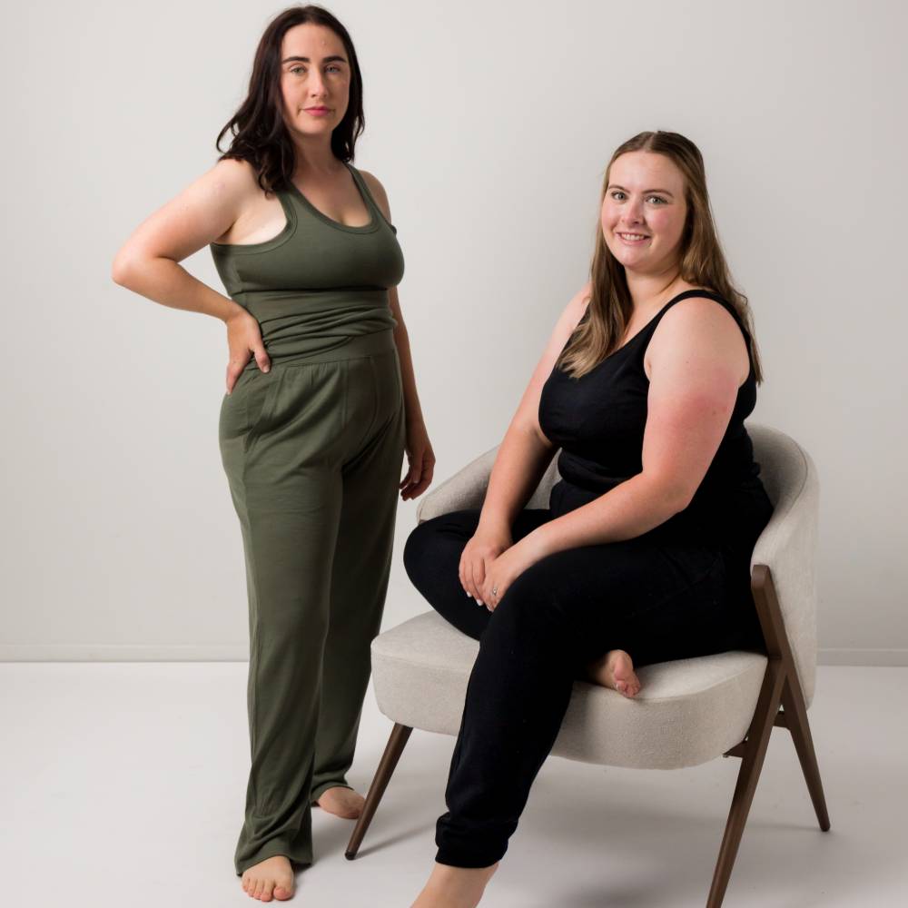 Two women modeling matching lounge sets in a studio setting. One woman stands barefoot wearing an olive green tank top and matching pants, while the other sits on a light beige armchair wearing a black tank top and black pants.