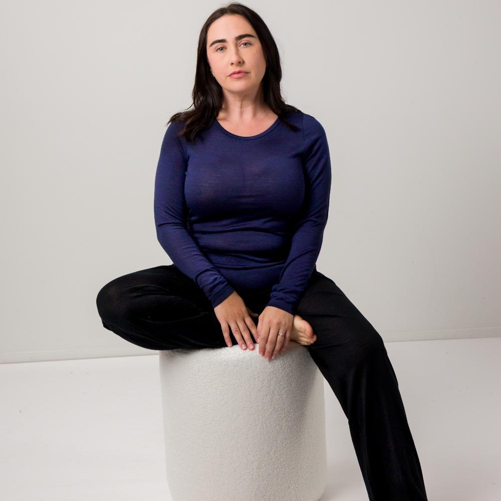 A woman sits cross-legged on a white cylindrical pedestal in a studio setting, wearing a dark navy long-sleeve top and black pants against a light gray background.