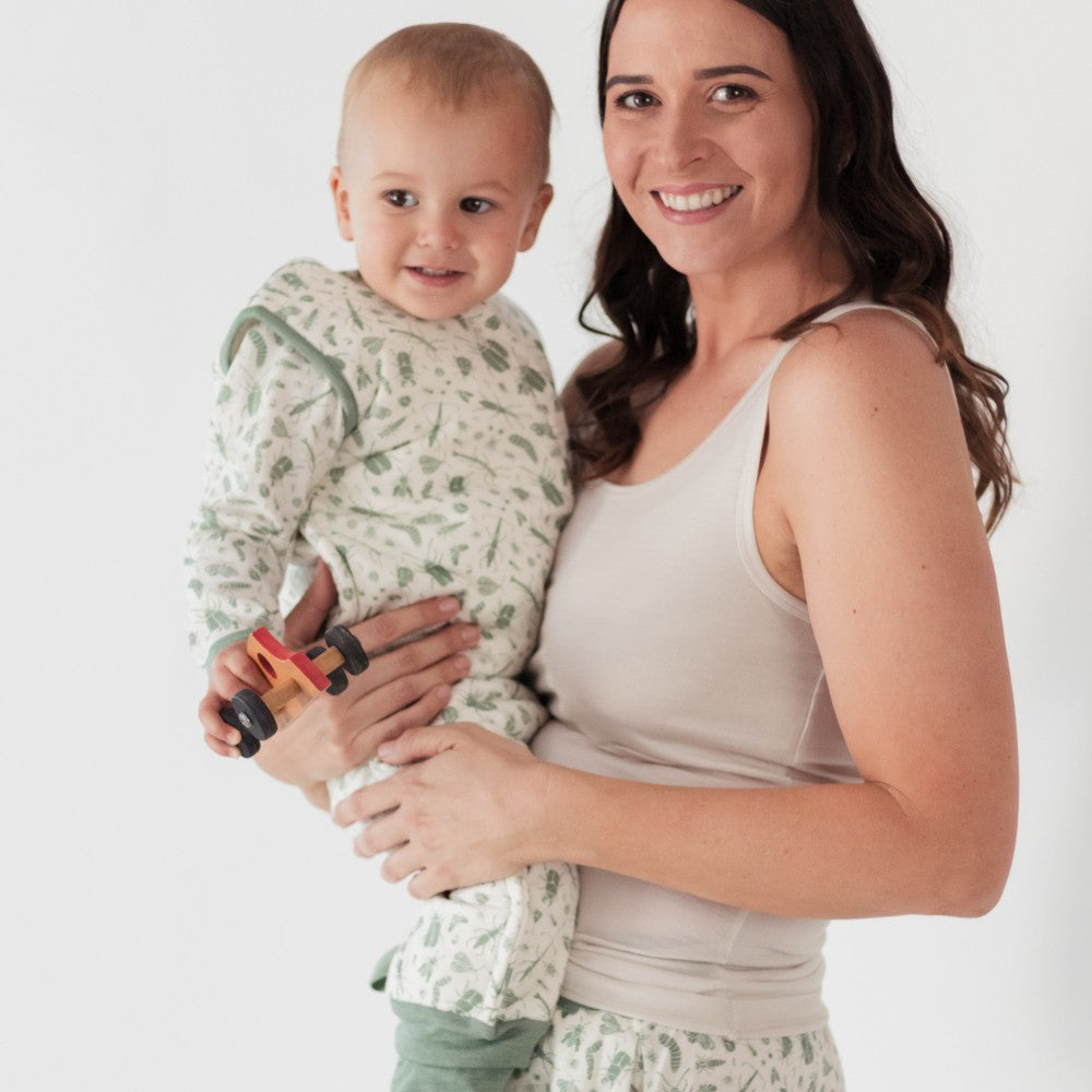 A smiling woman holds a baby wearing a light green and white patterned long-sleeve sleepsuit against a clean white studio background. The woman wears a white tank top.