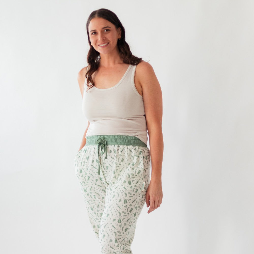 A woman modeling a pair of white pants with a green botanical pattern and a green elastic drawstring waist, paired with a white tank top, against a plain white background.