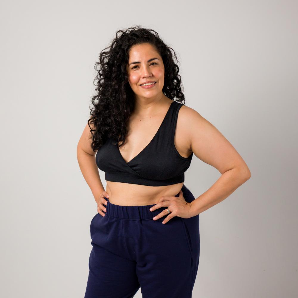 A woman with curly dark hair stands against a light gray studio background, wearing a black wrap-style sports bra and navy blue sweatpants, with her hands on her hips.