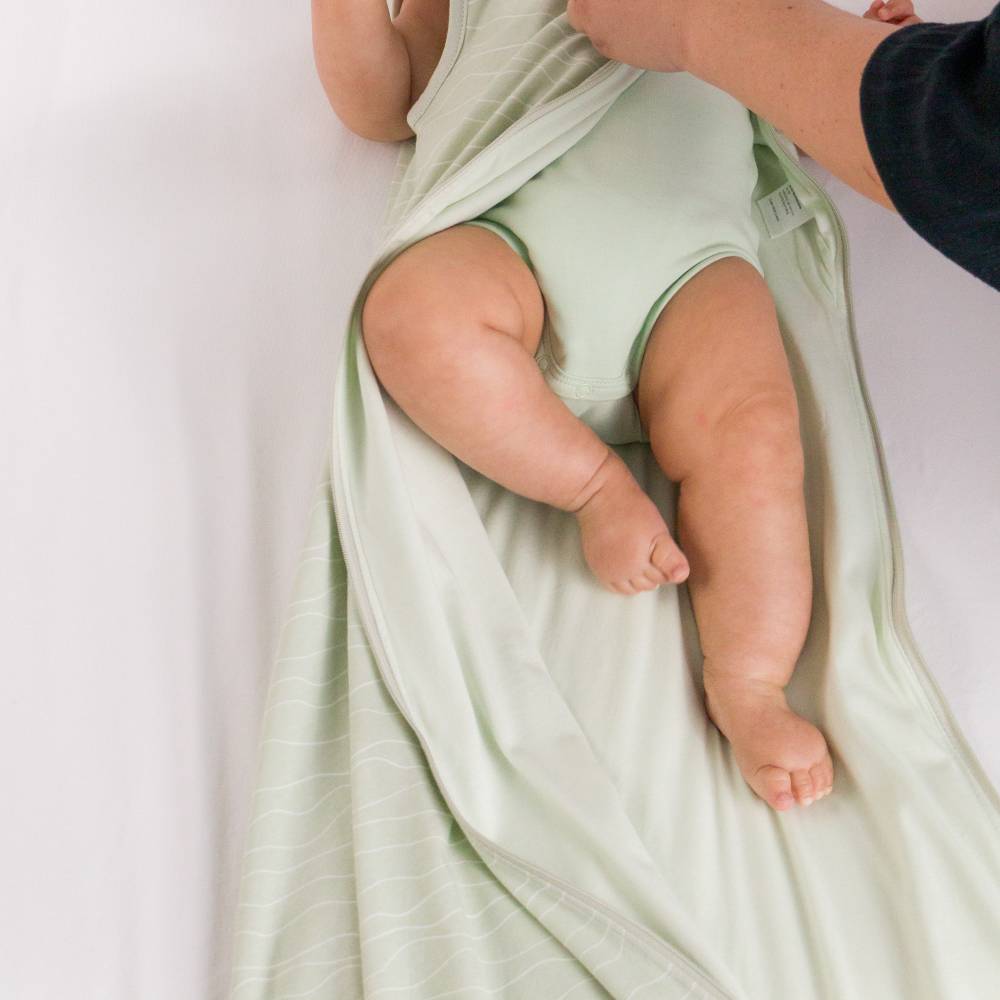 A baby wrapped in a pale sage green cotton sleeping bag wearing a matching light green bodysuit, with an adult's hand adjusting the swaddle.