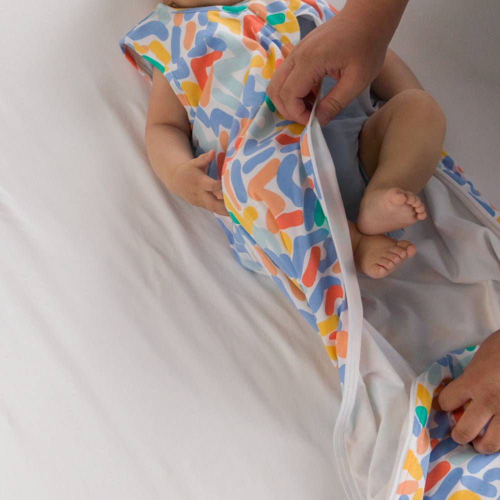 A baby wearing a colorful abstract print with an adult's hands helping to pull the front flap open over white bedding.