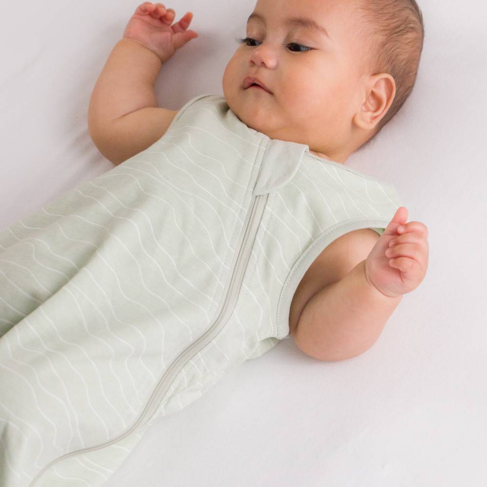 A baby lying on a white blanket wearing a pale sage green sleeveless sleep sack with a front zipper.