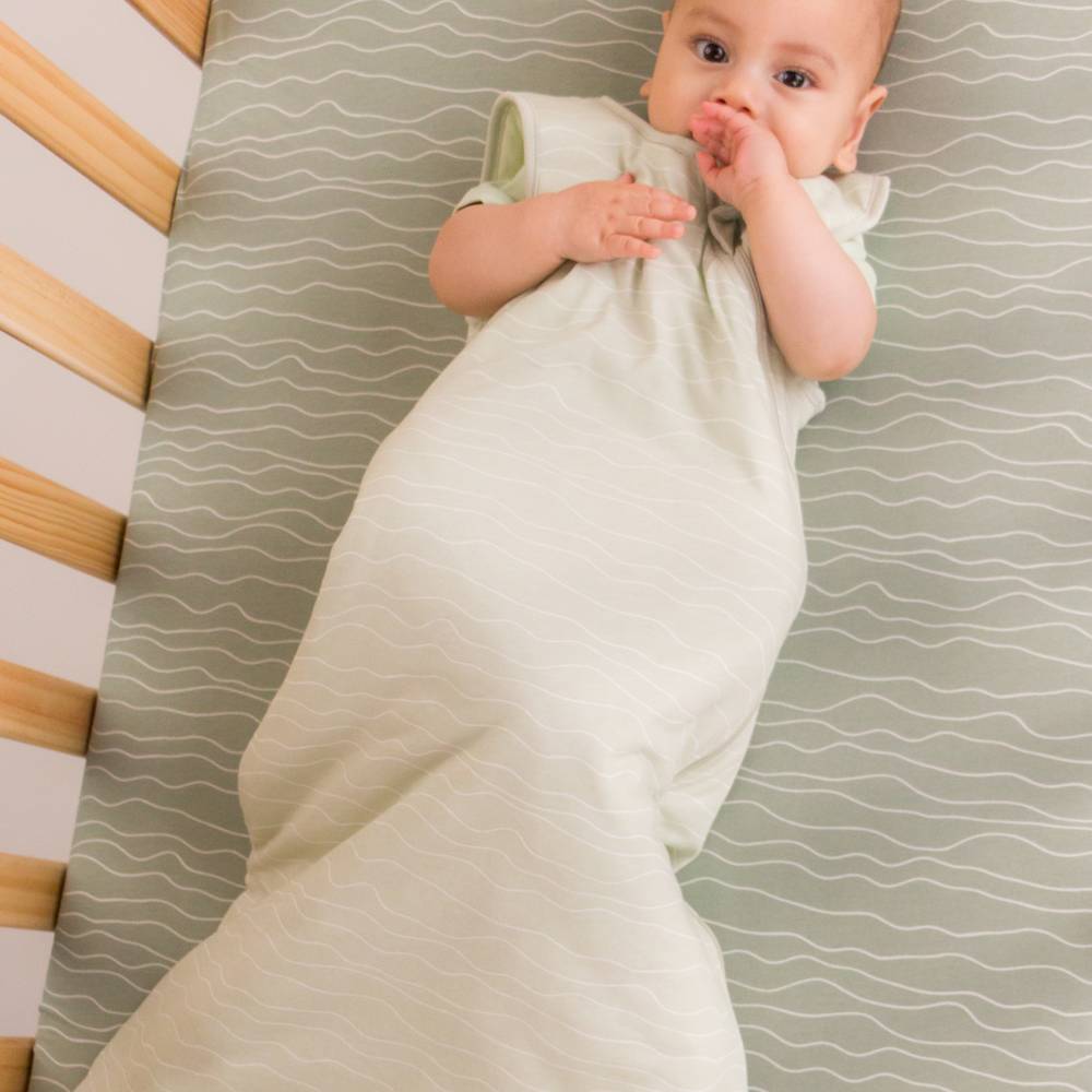 A baby lying on their back in a crib,  in a pale sage green sleep sack with no sleeves. The crib mattress has a matching sage green and white wavy pattern, and the wooden slats of the crib are visible on the left.