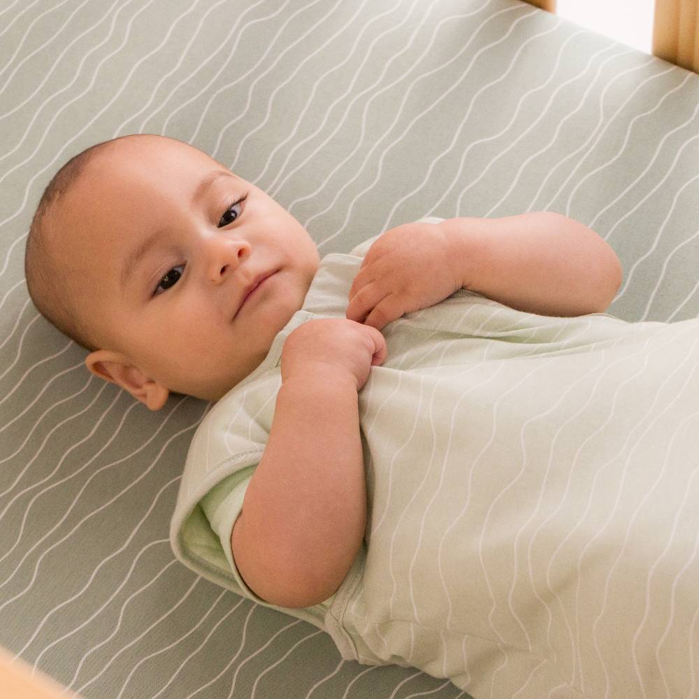 A baby lying on their back on a striped green cot mattress wearing a pale sage green short-sleeve bodysuit.