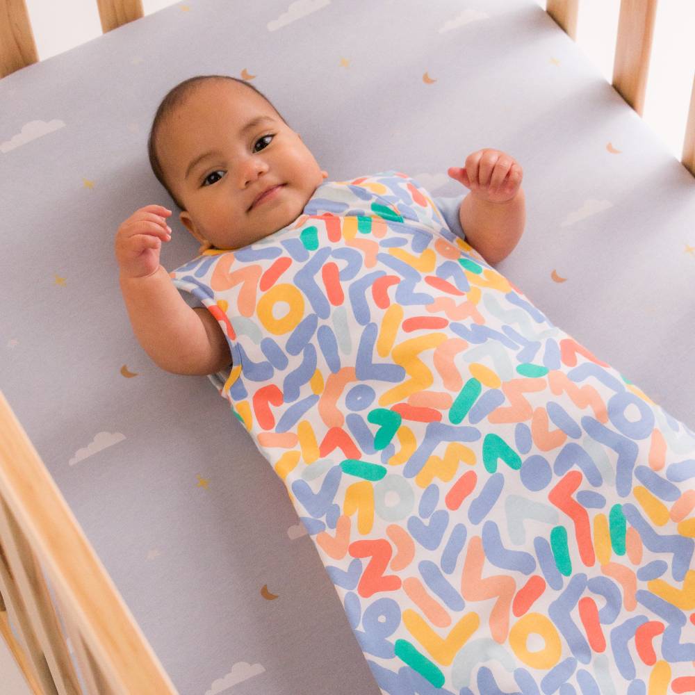 A baby lying on their back in a wooden crib, wearing a light blue sleep sack with a colorful abstract pattern of orange, yellow, green, and blue shapes. The crib mattress has a matching light blue sheet with small white clouds and gold crescent moons.