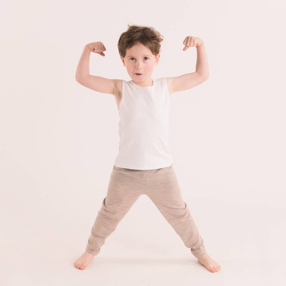 Child flexing muscles wearing a white singlet tank top and beige pants on a light background