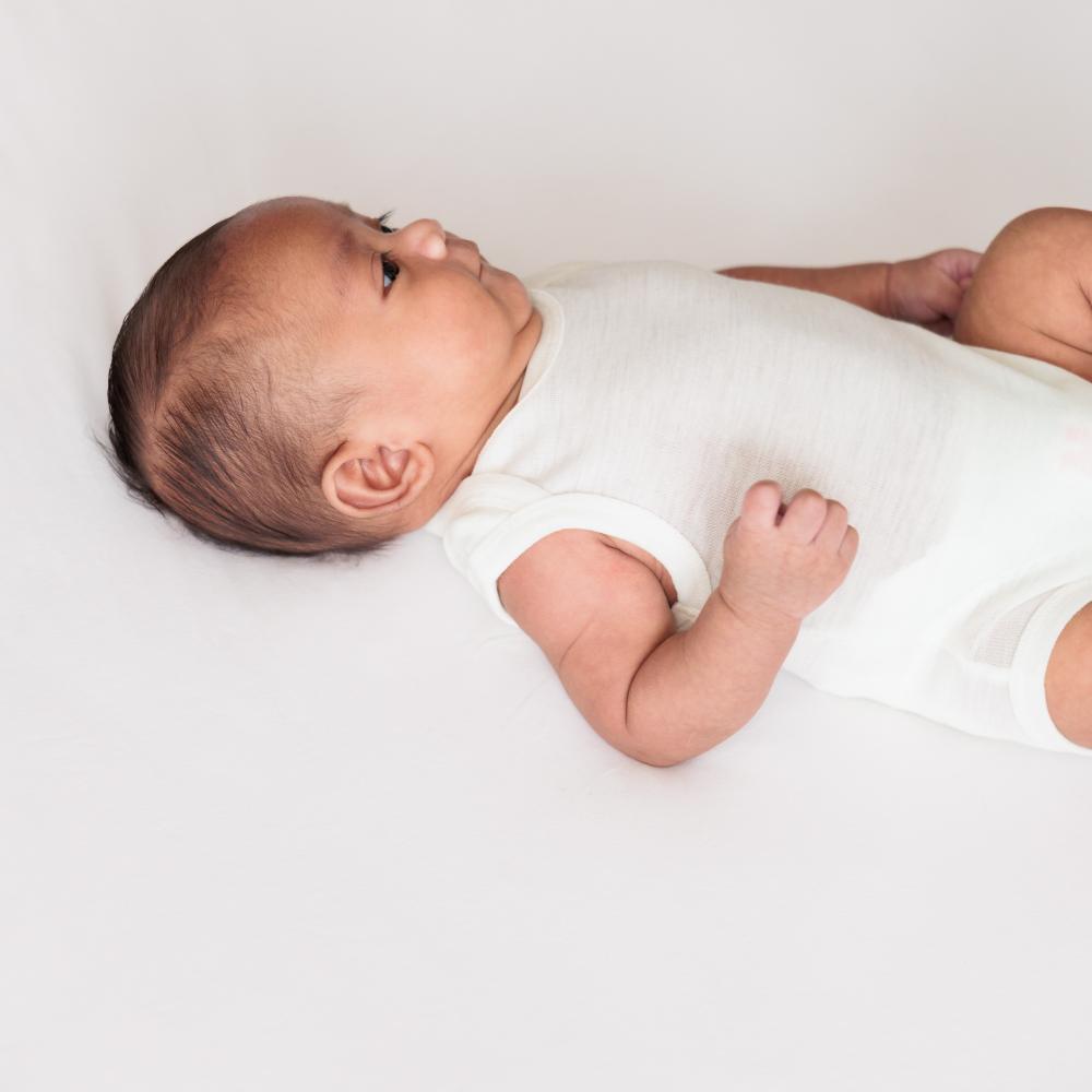 A baby lying on their side wearing a white merino wool singlet bodysuit, photographed against a clean white background.