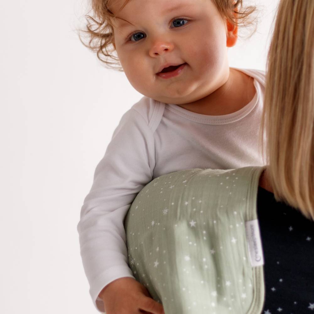 Baby in a white bodysuit held by a person wearing a green burp cloth with a visible the sleep store label.