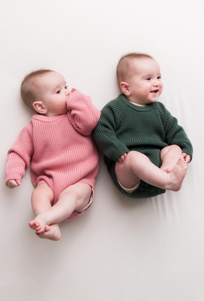 Two babies in pink and green knitted merino wool bodysuit rompers lying on a white surface