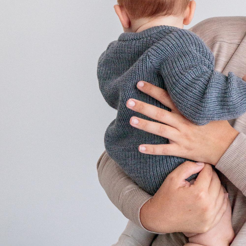Person holding a child wearing a gray merino knit bubblesuit against a plain background
