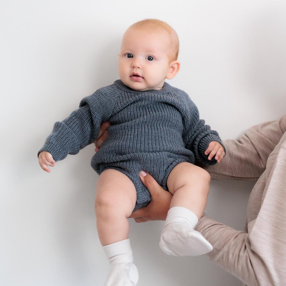 Baby in a grey knitted merino longsleeve bubblesuit outfit being held against a plain background