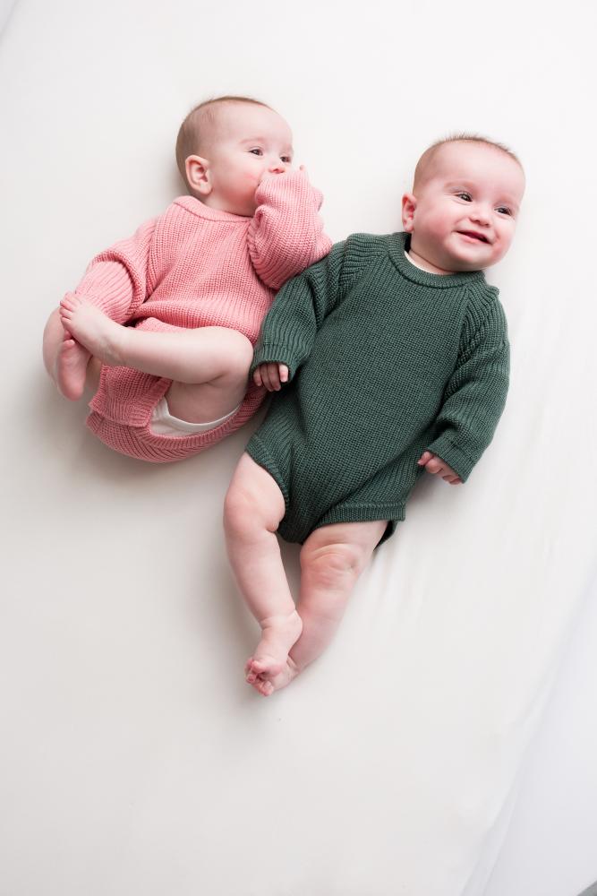 Two babies in pink and green knitted merino bubblesuit romper outfits lying on a white surface.