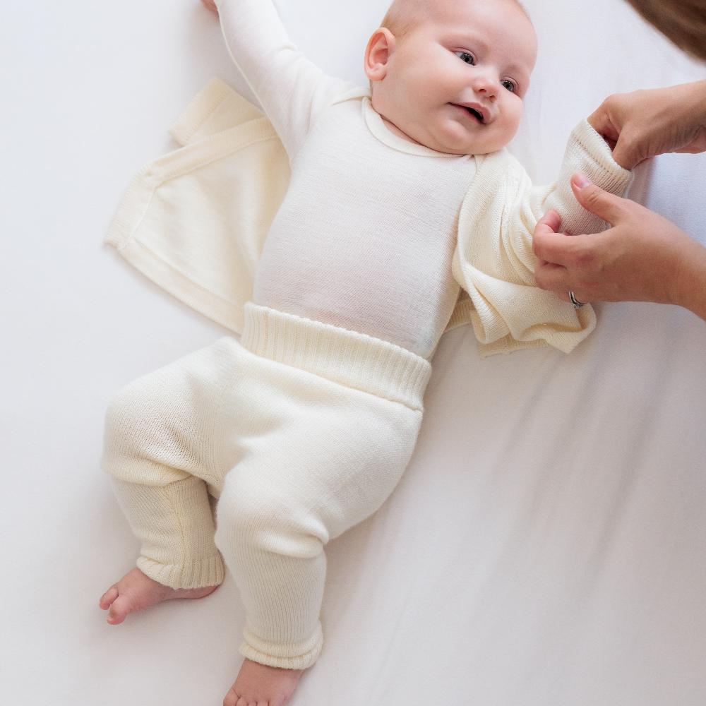 Baby in a cream-colored merino knitted outfit lying on a white surface with a person's hand holding