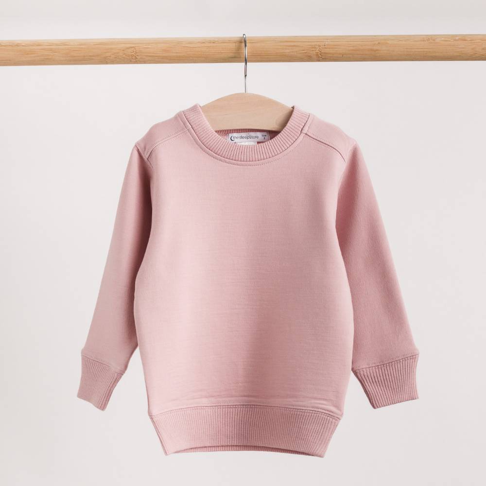 A light pink long-sleeve merino fleece/tencel crewneck sweater hanging on a wooden hanger from a wooden clothes rack, set against a plain off-white background.