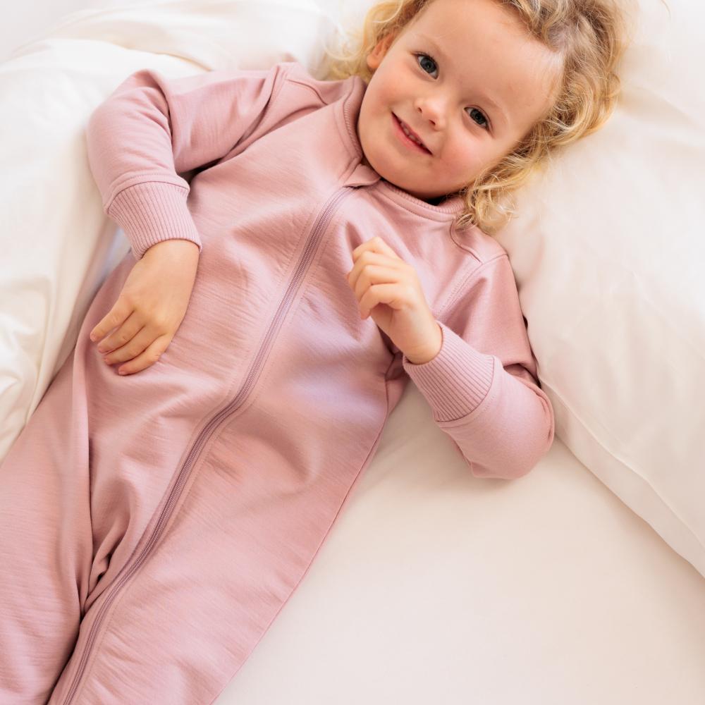 A smiling toddler lying on white bedding, wearing a dusty pink merino fleece long-sleeve zip-up sleeper.