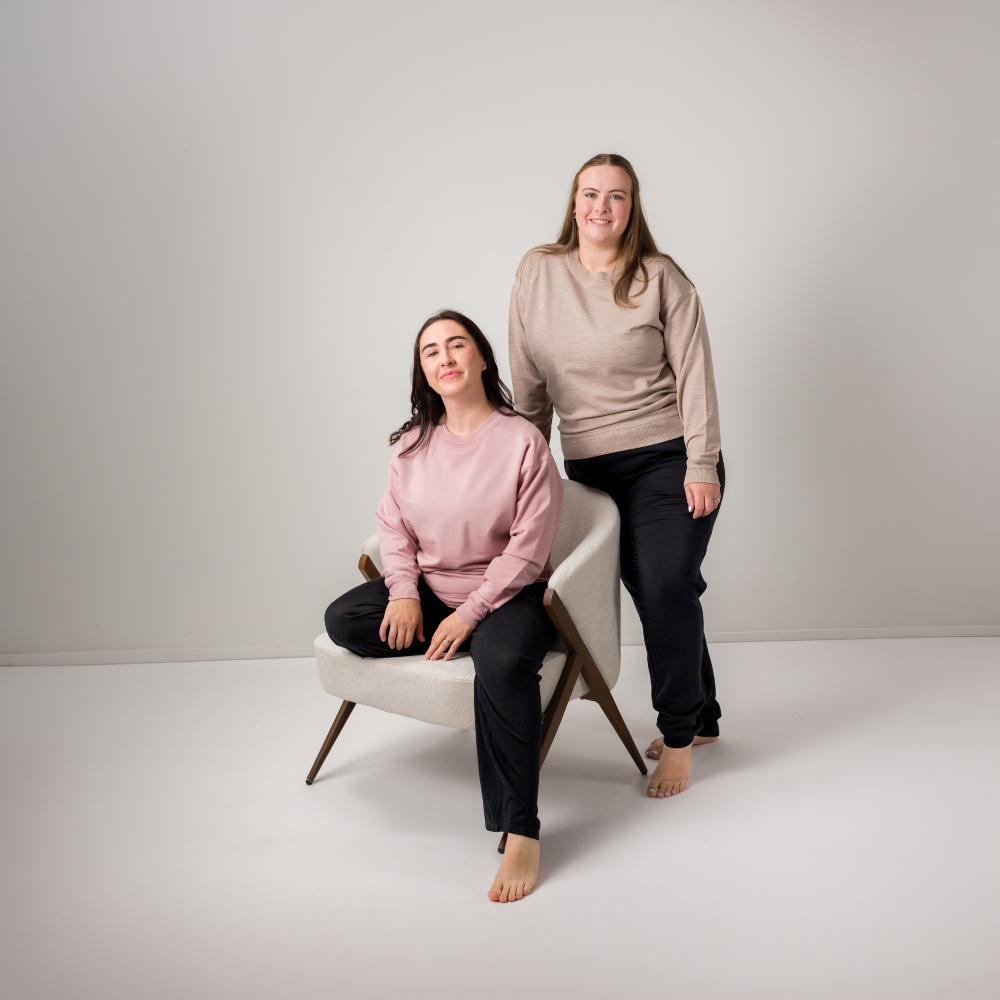 Two women modeling matching long-sleeve crewneck merino fleece  sweatshirts in a studio setting. One woman is seated on a modern light gray armchair wearing a dusty pink sweatshirt and black pants, while the other stands behind her wearing a light beige sweatshirt and black pants.