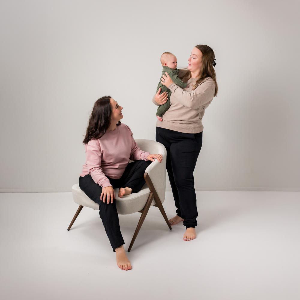 Two women in a minimalist studio setting, one seated on a white armchair in a pink top and black pants, one standing holding a baby in an olive green outfit, and another standing in a beige merino top and black pants.