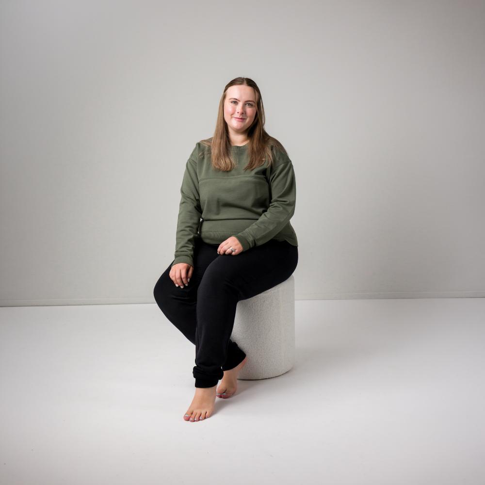 A woman sits barefoot on a cylindrical white stool in a studio setting, wearing an olive green long-sleeve top and black merino tencel jogger pants against a light gray background.